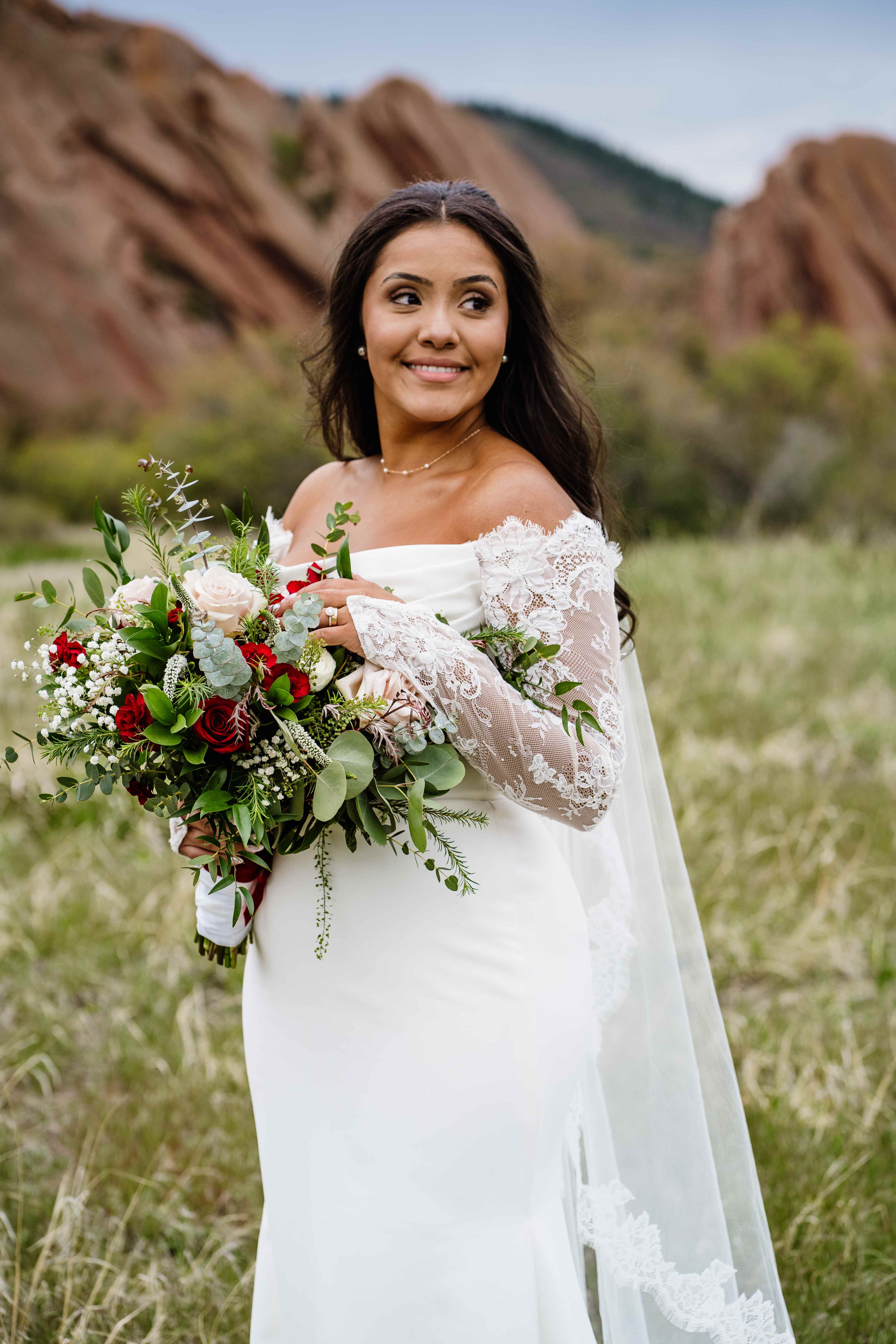 Bride with a warm sun-kissed glow in an off-the-shoulder lace gown holding a red rose bouquet, with Colorado's red rock formations behind her