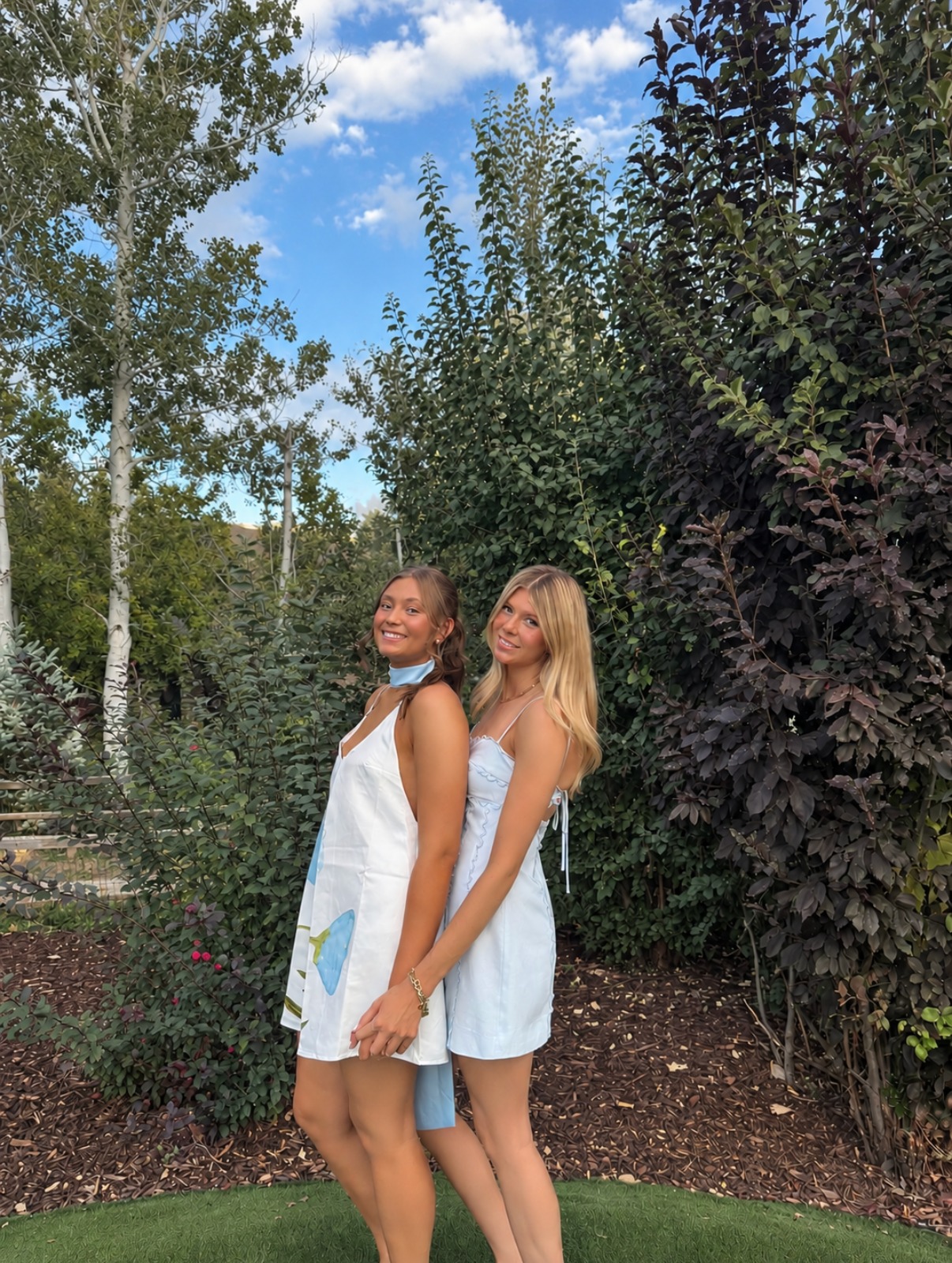 Two friends in white and light blue mini dresses posing back to back in a garden with sun-kissed homecoming spray tans