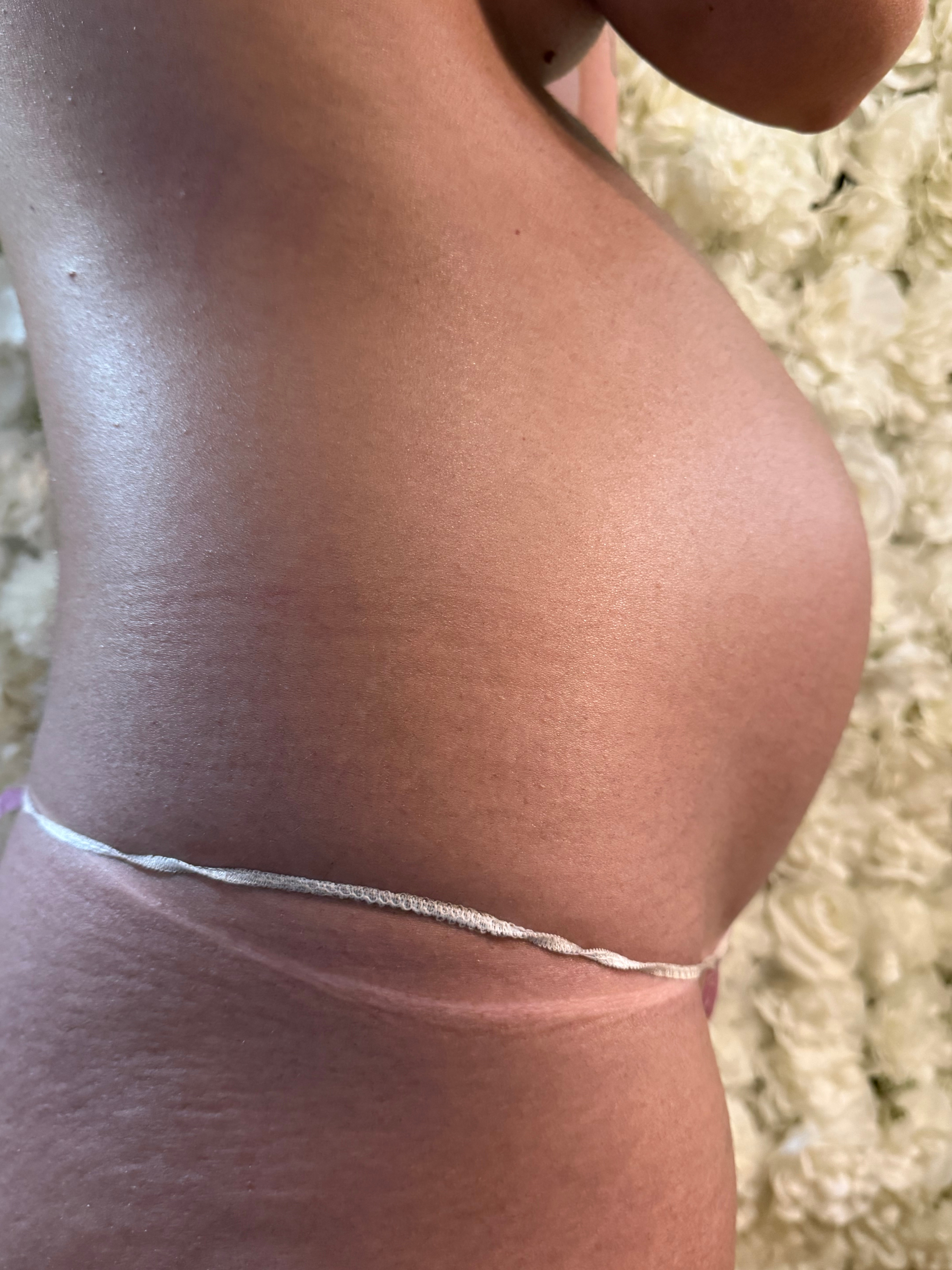 Side profile of a pregnant client's baby bump with a deep, even, streak-free bronze spray tan in front of a white floral wall