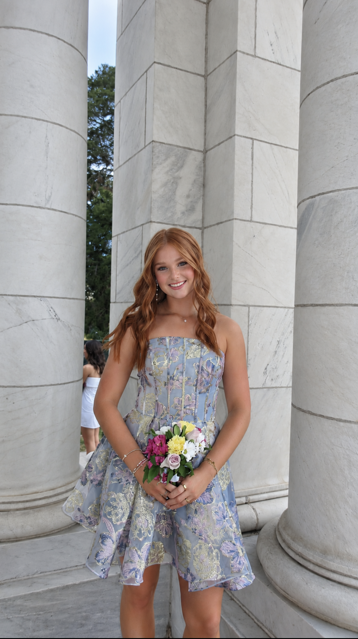 Redheaded client in a strapless blue floral homecoming dress holding a bouquet, glowing with a soft, natural spray tan between marble columns