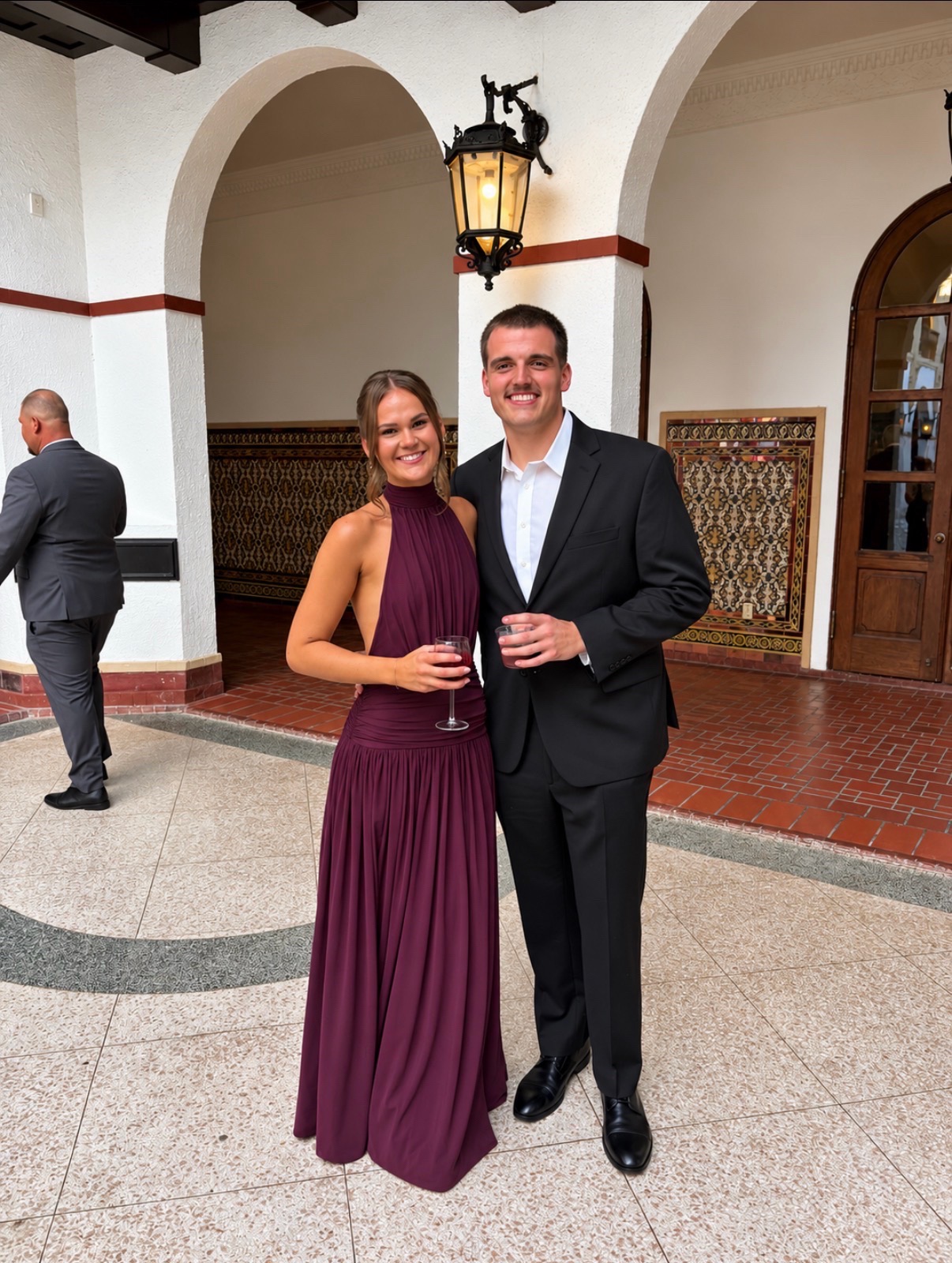 Wedding guest in a plum halter gown with a natural, radiant spray tan posing with her date beneath Spanish-style arches