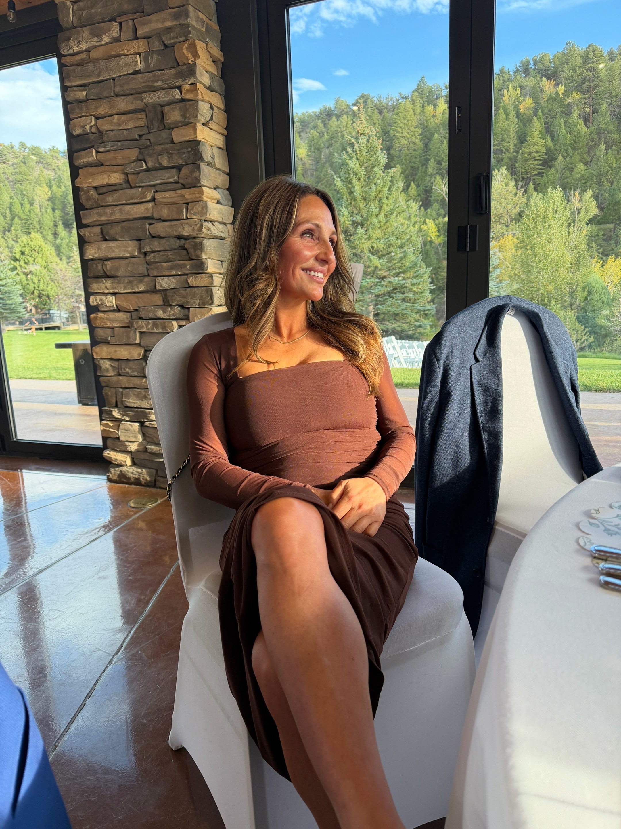 Smiling wedding guest in a chocolate brown dress with a radiant, natural spray tan at a mountain venue